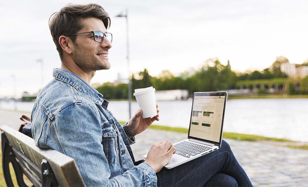 Chico sentado en un banco mientras bebe un café y mira el ordenador tranquilamente